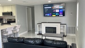 Una sala moderna con un sofá de cuero negro, un televisor de pantalla plana sobre una chimenea, y una cocina al fondo.