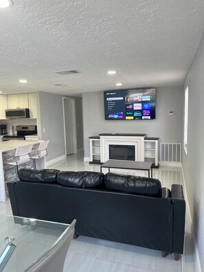 A modern living room with a black leather sofa, a flat-screen TV mounted above a fireplace, and a kitchen area in the background. - 2-bedroom house in pleasant Port Richey with cool AC (Port Richey)