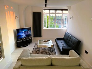 A modern living room with a black leather sofa, a white coffee table, a flat-screen TV, and a large window with a view of greenery. - Stylish 2Bed Flat-Fast WiFi-Close to Shops, Restaurants & Train Station-Sleeps 5 (Croydon)