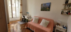 A room with a wooden floor, a white desk with a chair, a pink sofa with white cushions, a painting on the wall, and a small bedside table with a lamp and tissue box. - Air-conditioned apartment in the heart of town, just 7' from the beach! (Nice)
