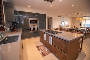 A modern kitchen with a central island, a dining area with a view, and a living space with a sofa and table. - Tau - Condo Kumar Inside the Gates (Punta Mita)