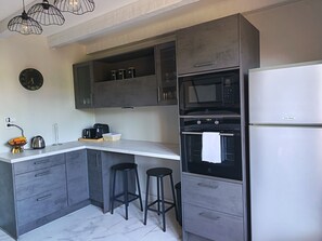 Una cocina moderna con armarios grises, encimera blanca, microondas negro, refrigerador de acero inoxidable y dos taburetes negros.