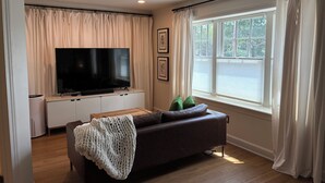 Ein Wohnzimmer mit Flachbildfernseher, einem braunen Sofa, einer weißen Decke und einem Fenster mit Vorhängen.