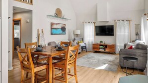 Un vaste espace de vie comprenant une table à manger, des chaises en bois, un canapé gris, un téléviseur à écran plat et un escalier.