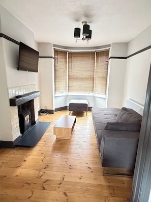 A living room with a fireplace, a sofa, a coffee table, and a window with blinds. - 3 Bed House Contractor Friendly Long Stays Welcome (Bedford)