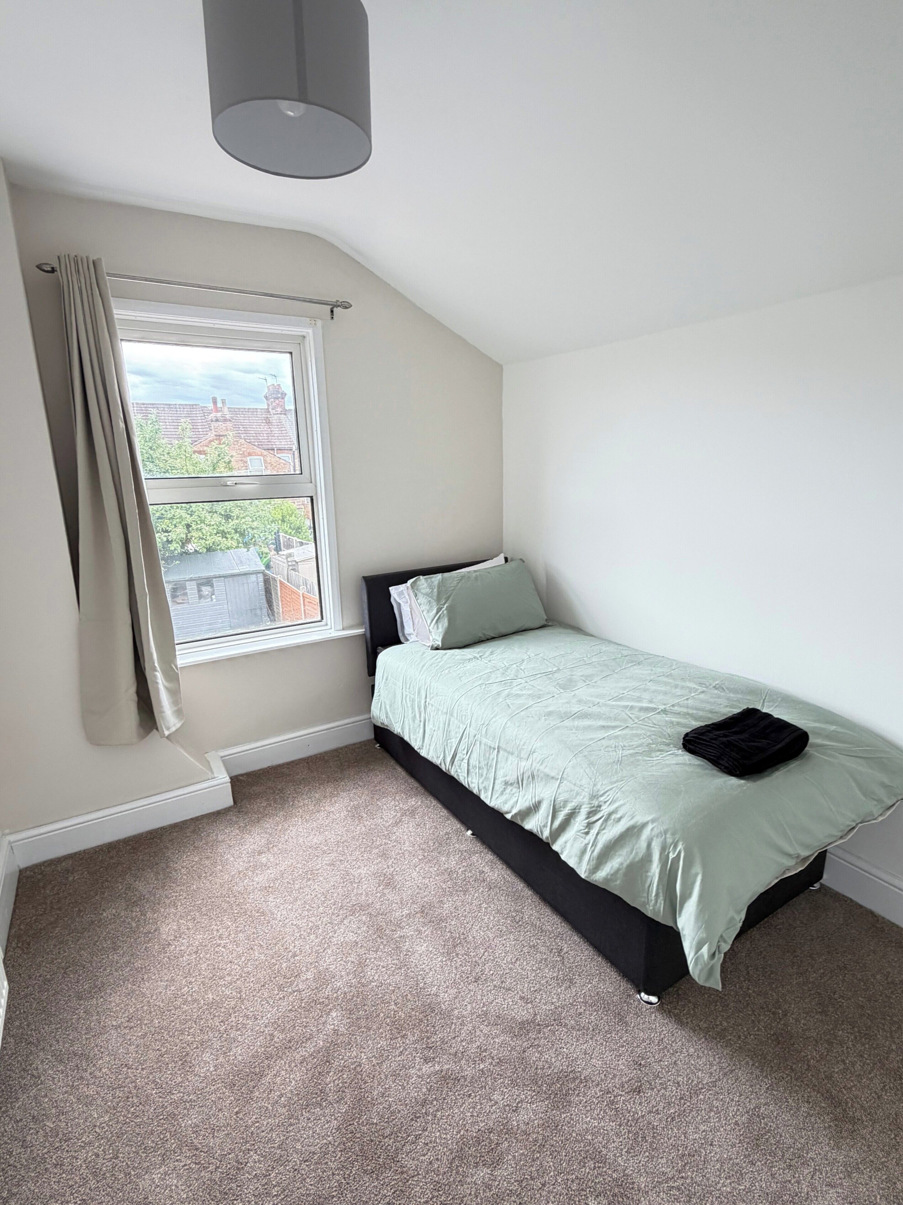 Una camera da letto con un letto singolo, una finestra con tende e vista su un quartiere residenziale.