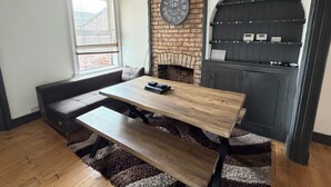 A cozy room with a brick fireplace, a wooden table, a bench, and a shelving unit with electronic devices. - 3 Bed House Contractor Friendly Long Stays Welcome (Bedford)