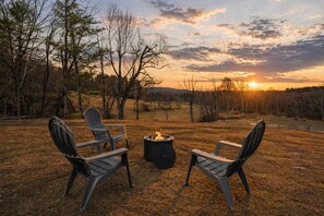 A fire pit with two Adirondack chairs facing it, set in a grassy field with trees in the background. - Fire Pit, BBQ & Room for Everyone, Spacious 4BR/3BA in Hendersonville (Hendersonville)