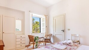 Quarto com cama, escrivaninha, duas cadeiras, cômoda e janela com vista para árvores.