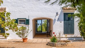 Um edifício branco com porta e janelas de madeira, um banco na frente e plantas em vasos.