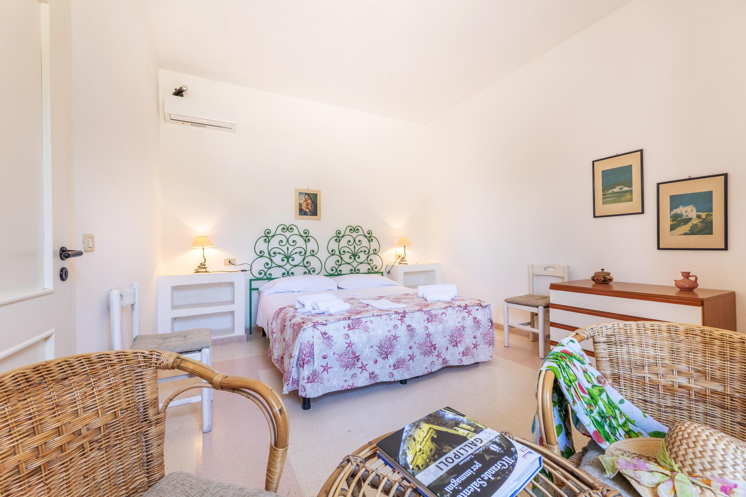 Una camera da letto con un letto, due comodini, un tavolino, una sedia in vimini e un condizionatore d'aria a parete.