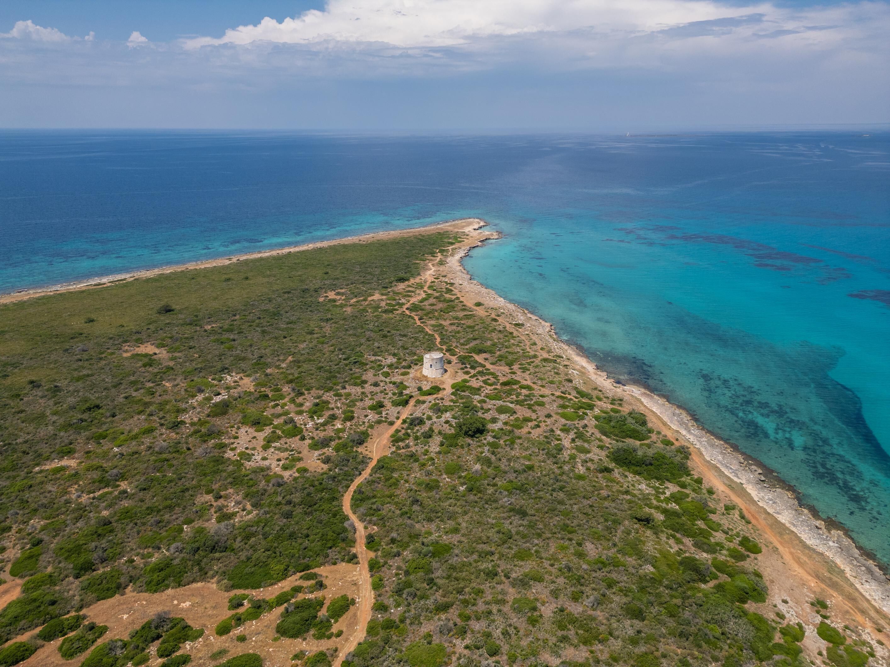 Vista aérea de uma área costeira com um farol, uma praia estreita e um caminho que leva ao farol.