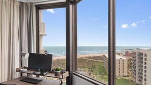 Ein Zimmer mit großem Fenster mit Blick auf einen Strand, einem Schreibtisch mit Computer und einer Ansicht eines Gebäudes gegenüber dem Wasser.