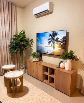 A living room with a TV, wooden cabinet, two stools, and potted plants. - Milyleo Home, Your Caribbean Hideaway – Private Jacuzzi & Tropical Comfort (Punta Cana)