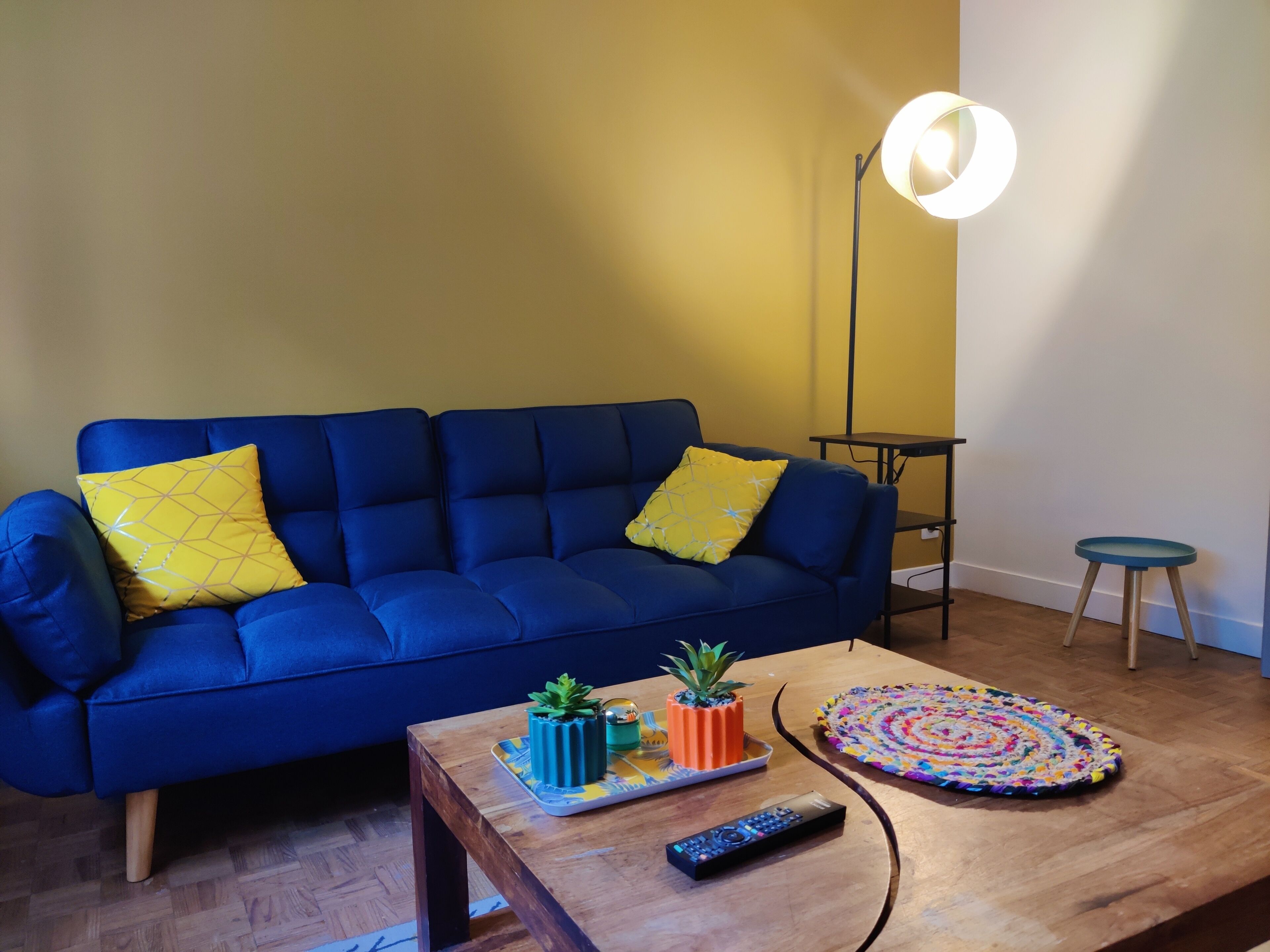 A blue sofa with yellow pillows, a wooden coffee table with a remote control, a small potted plant, and a colorful rug.