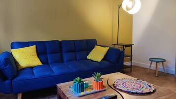 Ein blaues Sofa mit gelben Kissen, ein Couchtisch aus Holz mit einer Fernbedienung, ein kleiner Topfpflanze und ein farbenfroher Teppich.
