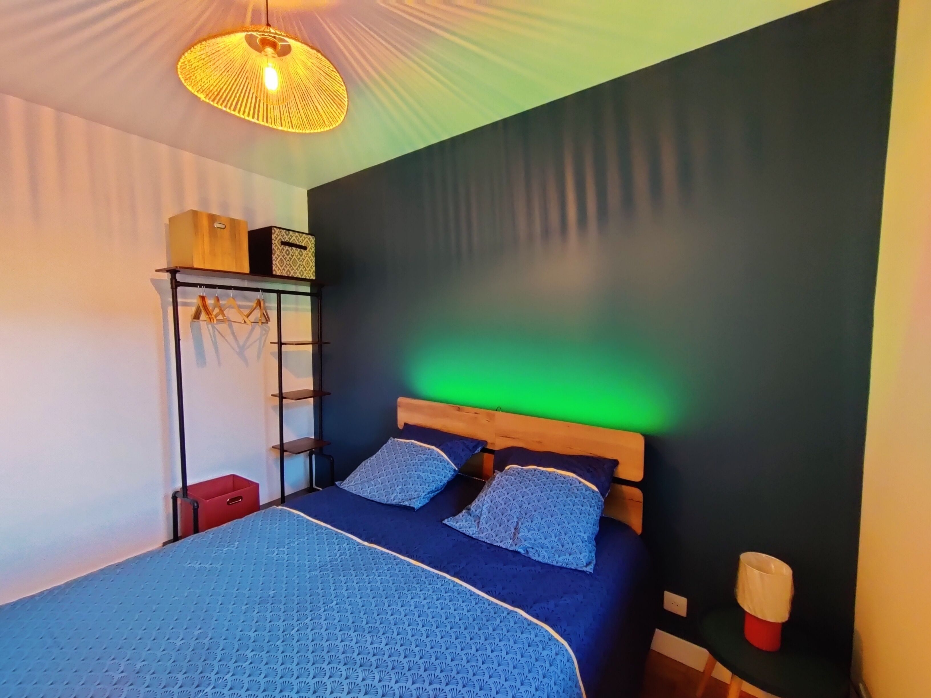 Una habitación con cama, lámpara colgante, estante con cajas y pared verde.