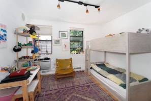 Ein Kinderzimmer mit Etagenbetten, einem Schreibtisch, Regalen mit Spielzeug und einem Fenster mit Blick auf Bäume.