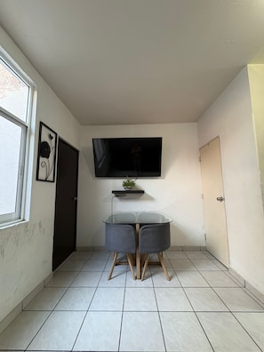 Una stanza piccola con un tavolino in vetro, due sedie, una TV fissata al muro, una porta e una finestra.