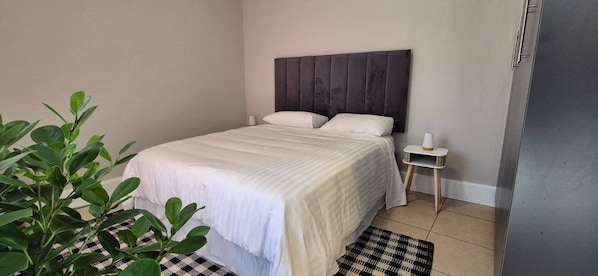 A bedroom with a bed, bedside table, lamp, and a plant. - Cozy 2 Bedroom Cottage (Johannesburg)