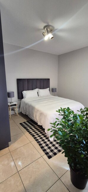 Un dormitorio con cama, ventilador de techo, mesita de noche con lámpara, planta en maceta y alfombra a cuadros.