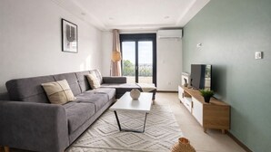 Una sala de estar moderna con un sofá gris, una mesa de centro, un televisor sobre un mueble de madera, y un balcón con vistas.