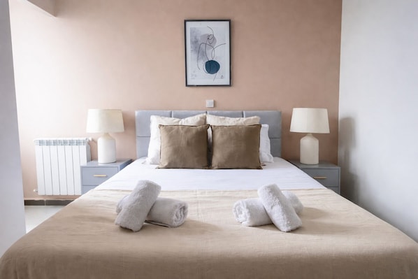 Habitación de hotel con una cama, dos mesitas de noche, un radiador y una obra de arte enmarcada en la pared.