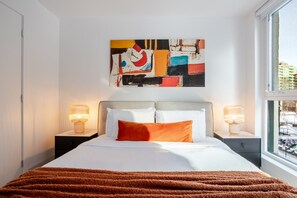 Ein Hotelzimmer mit Bett, zwei Nachttischlampen, einem großen abstrakten Gemälde und einem Fenster mit Blick.