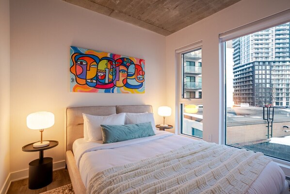Ein modernes Schlafzimmer mit einem großen Bett, zwei Nachttischlampen, einem farbenfrohen abstrakten Gemälde und Blick auf eine Stadtlandschaft durch ein großes Fenster.