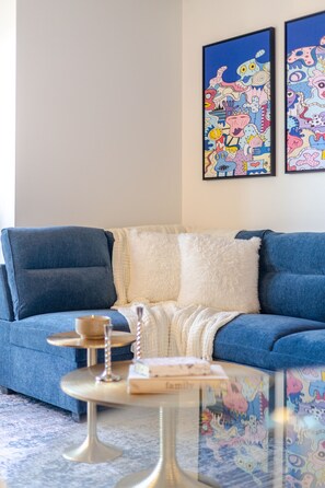 Un canapé bleu agrémenté de coussins blancs et moelleux, une petite table ronde avec une bougie, et deux tableaux abstraits aux couleurs vives accrochés au mur.