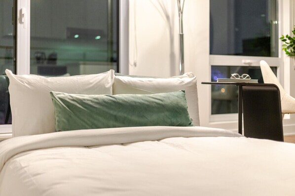 A bed with white bedding and a green pillow, a bedside table with glasses, and a window with a view of the outside. - Urban Oasis with Parking (Montreal)
