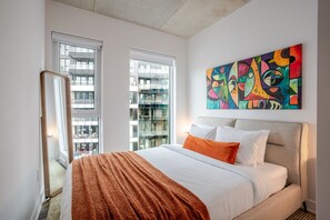Habitación de hotel moderna con una cama grande, un cuadro abstracto de colores vivos y vistas a la ciudad a través de amplias ventanas.