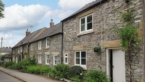 A row of stone houses with white doors and windows, a paved road, and greenery along the sidewalk.