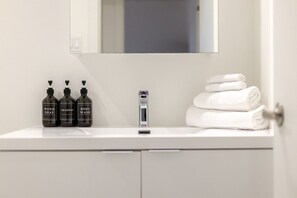 Ein Badezimmer mit weißem Waschtisch, drei Flaschen Handseife, Duschgel und Shampoo, einem Rasierer und einem Stapel weißer Handtücher.