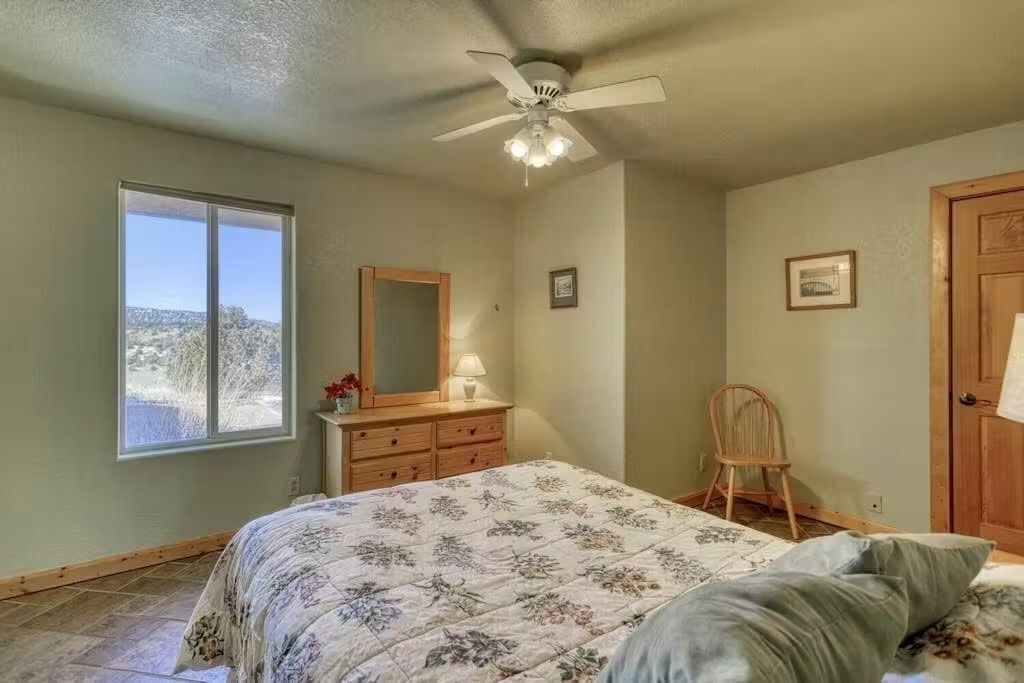 A bedroom with a bed, a ceiling fan, a dresser, a chair, and a window with a view.