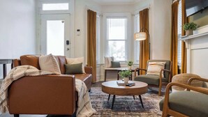 Ett vardagsrum med en brun soffa, ett runt soffbord och en stol. Det finns fönster med gardiner, en växt på ett sidobord och en hängande lampa.