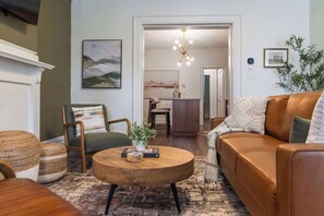 A living room with a brown leather sofa, a round wooden coffee table, and a green armchair. There is a painting on the wall and a plant on the floor. - Hot Tub - Sleeps 9 - Near OSU (Columbus)