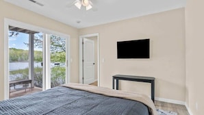 Une chambre à coucher comprenant un lit, un téléviseur, un ventilateur de plafond et une vue sur une étendue d’eau, visible à travers de grandes fenêtres.