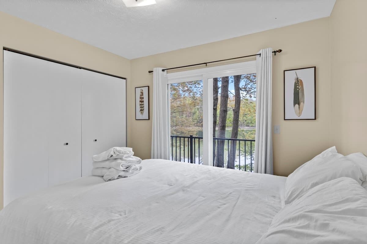 A bedroom with a bed, a window with a view of trees, a door, and a framed picture on the wall.