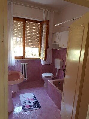 Ein Badezimmer mit Toilette, Waschbecken und Badewanne. Es gibt ein Fenster mit Jalousien, eine Heizung und eine Tür.