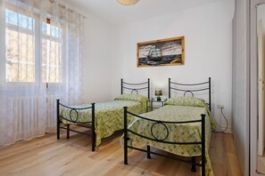 Ein Zimmer mit zwei Betten mit Metallrahmen, einem Gemälde eines Schiffes an der Wand und einem Fenster mit transparenten Vorhängen.