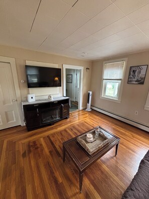 Un salon avec un plancher en bois, un téléviseur posé sur un meuble, une table basse et une porte donnant sur une autre pièce.
