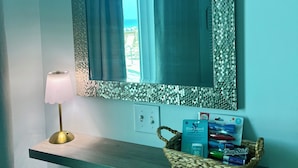 A mirror with a textured frame, a lamp, and a basket of toiletries on a shelf. - Oceanfront Condo, Gated Entry, Pool, King Bed, <100 Steps to Sand.  (Pensacola)
