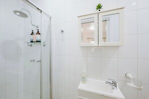 A bathroom with a glass-enclosed shower, a white sink with a mirror above it, and a small shelf with toiletries. - Claret Cottage Walk into Mudgees Historic Heart (Mudgee)