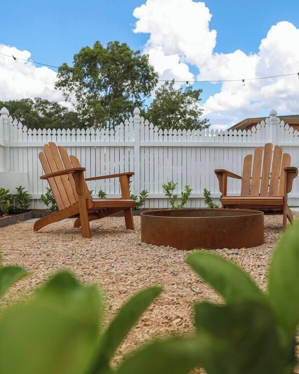 Zwei Adirondack-Stühle, eine Feuerstelle aus Metall und ein weißer Zierzaun in einem Garten.