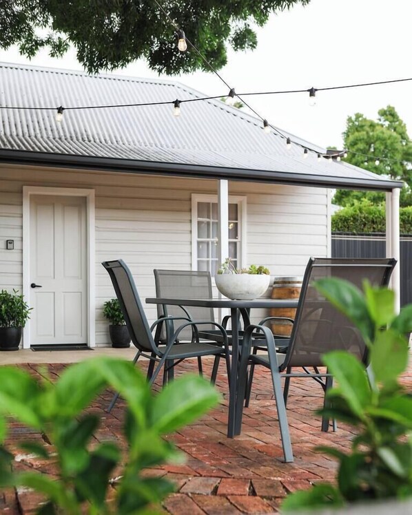 A house with a white exterior, a grey roof, and a brick patio. There is a table with chairs and a potted plant.