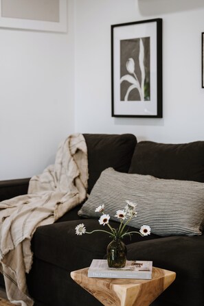 Een woonkamer met een donkere bank, een gestreept kussen, een vaas met witte bloemen en een boek getiteld 'The Quiet Home' op een houten bijzettafeltje.