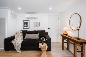 A modern living room with a dark sofa, a coffee table, a wooden side table with a lamp, and a wall-mounted air conditioner. - Luxe Loft Contemporary Comfort by Mudgees CBD (Mudgee)