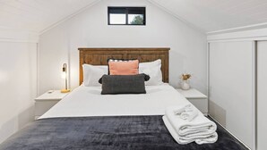 Un dormitorio con cabecera de madera, cama con ropa de cama blanca, mesita de noche con lámpara y una ventana pequeña.
