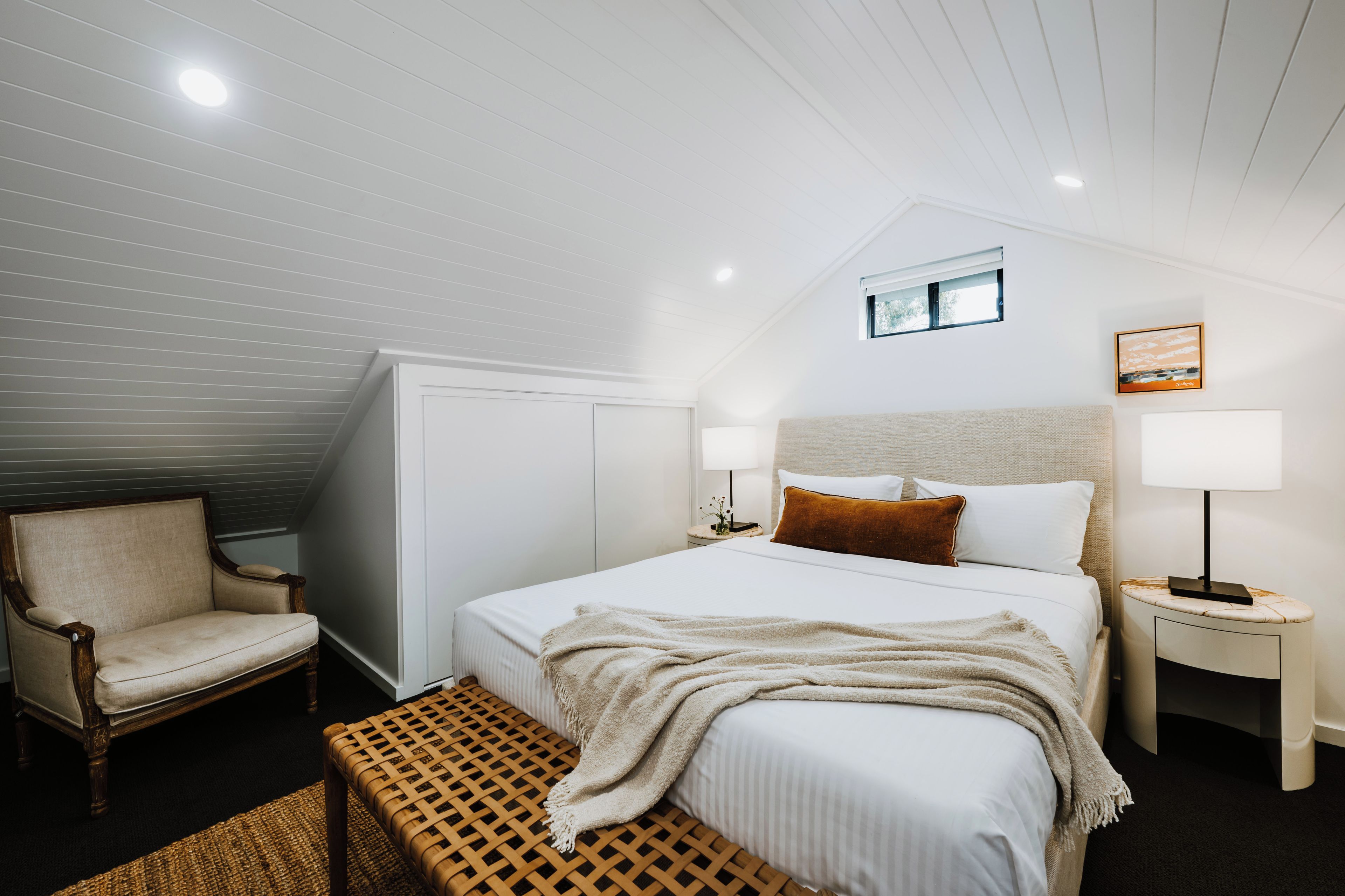 A bedroom with a bed, a chair, a bedside table, a lamp, and a window.